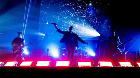 Three members of Coldplace performing on stage/ Two are holding guitars and the man in the middle is holding a microphone. The stage lights are blue and red.