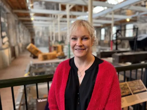 A woman with blonde hair, wearing a red cardigan and black top. She is standing in a museum with exhibits visible behind her