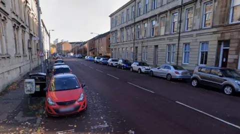 Google A general view of a street, with cars parked along either side of the road. The street is lined by three-storey stone buildings, white autumnal leaves scattered on the road. 