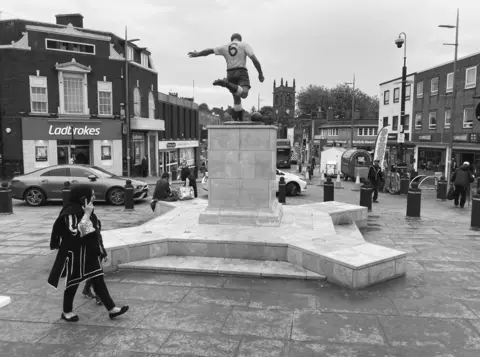 Chris Moss A statue, depicting Duncan Edwards in action, on top of a plinth is in the middle of the black-and-white photo, with buildings to the left and right of it on the image. One person is walking in front of the statue, while a few others are in the background. 