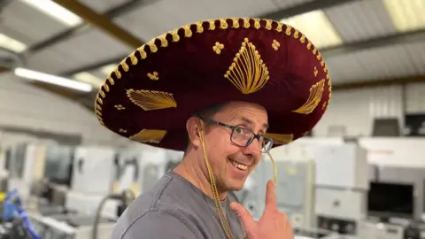 Daniel Thompson gives a slight side profile smiling and looking at the camera. He is wearing a Mexican-style large hat, covered in deep red velvet with gold decorative thread patterned onto it. A warehouse of blurred appliances in the background.