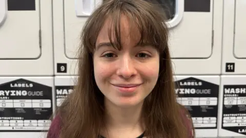BBC/Alice Lilley Ellie May is a first-year history student. In this image she is smiling in front of the washing machines in a laundry room on campus at the University of York. Ellie May is wearing a stripey red top and has straight brown hair with a fringe. 