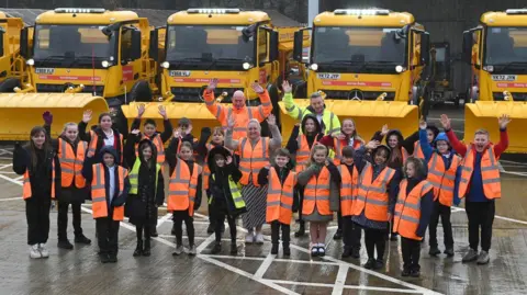 Sunderland City Council A group of school children wearing hi-vis jackets stand are waving at the camera. They are standing in front of a row of yellow gritters, which display their new names on red banners.