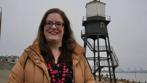 John Fairhall/BBC Jessica Bryan from Tendring District Council, standing in front of one of the Dovercourt lighthouses. She is wearing dark-rimmed glasses, a brown coat, a dark coloured floral top and a red beaded necklace.