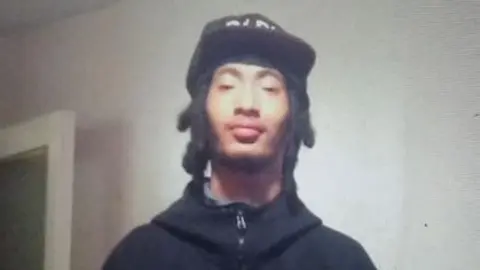 FAMILY HANDOUT Kalvin Taylor looks directly at the camera with a serious expression, he is wearing a black zip-up hoodie and black cap. 