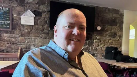 George Campbell. He is bald and wearing a grey shirt. He is sitting in the pub, which has brick walls and wooden tables behind him.