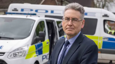 A man with grey hair and glasses in a navy suit with a blue shirt and blue tie, standing in front of two parked police vehicles