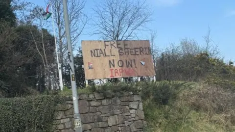 BBC A billboard that says FREE NIALL SHEERIN NOW! IRPWA It is on top of a grassy bank with a stone wall and lampost at the front. 