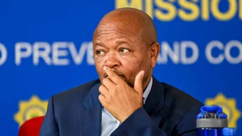 Police minister Senzo Mchunu, dressed in a dark blue suit and striped shirt, is seen holding his hand to his face during a briefing held by the police