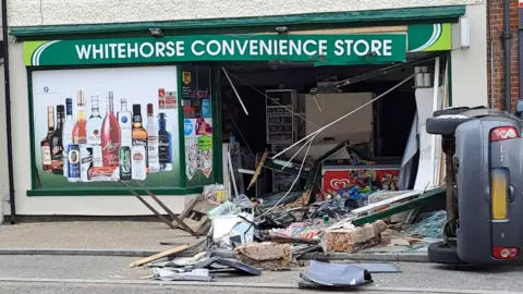 The front of a shop that has been severely damaged, with bricks, wood and metal lying on the ground. Fridges and shelves can be seen inside the shop. A window sign showing bottles on it is on the left and a grey car lies on its side to the right. 