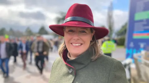 BBC Gemma Steve wearing a wine-red hat and green coat with racegoers walking behind her.