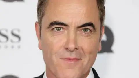Getty Images A man with black hair and black eyebrows is attending a red carpet event in a tuxedo. A number of blurred sponsors can be seen in the background wall.