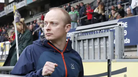 Getty Images A man with short fair hair, wearing a blue zipped up jacket in a football stand.