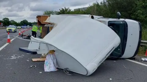A motorhome is on its side on the road. Police officers surround the car and much debris is on the road.