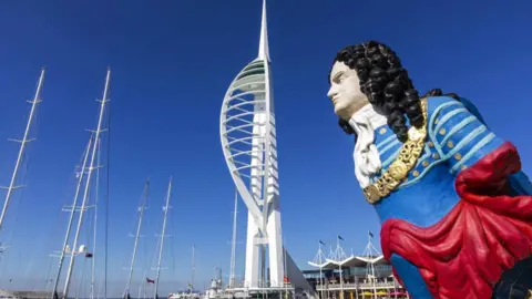 Getty Images The Spinnaker Tower, an iconic tall structure in the shape of a ship's sail which is painted white. In the foreground is the statue of a maritime ship's figurehead of a sailor, who has black curly hair and is wearing a blue nautical uniform.