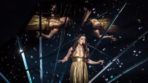 Getty Images Lucy, with long brown hair, is wearing a full-length gold dress and singing into a microphone on a stand. Behind her are mirrors, with reflections of herself, and sparkly lights.