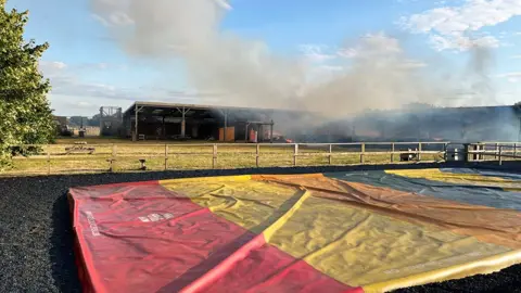 Smoke rising from burning building in the distance. It is an open-fronted wood structure in a field. There is a multi-coloured inflatable bounce pillow, deflated, laying on the ground in front of the barn.