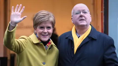 Nicola Sturgeon in a yellow jacket stands next to Peter Murrell