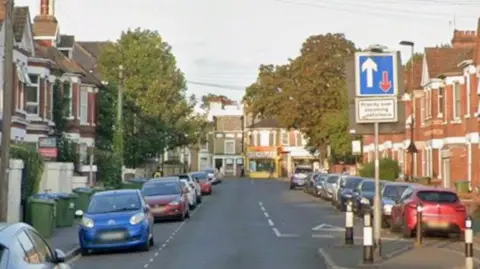 A residential road with cars parked on either side. Large trees can be seen at the far end of the street. 