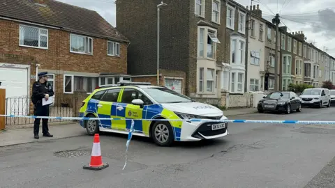 A police officer stands next to a patrol car on a cordoned off street. 
