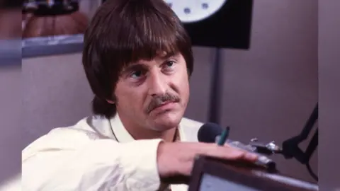 A vintage image of a brown haired man with a 1970s-era bowl haircut and moustache facing a microphone. 