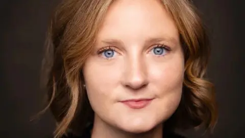 A woman's face, with short blonde hair and bright blue eyes. She is looking the camera. The background is black. 