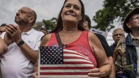 A woman, smiling and holding an American flag, stands in a crowd. She wears a red top.