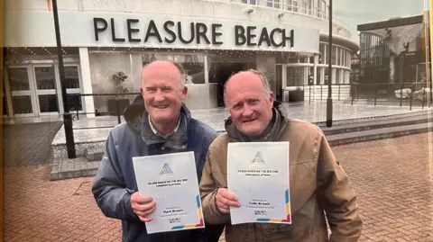 Handout Two men with silver hair smile at the camera while holding up white certificates which says they have ridden the Big One rollercoaster 20,000 times.