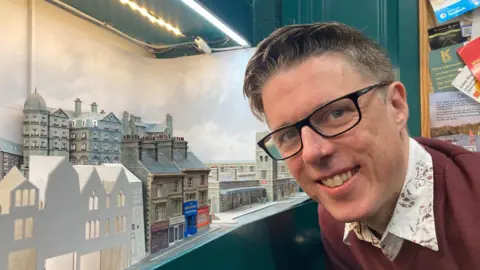 A man in glasses and shirt and maroon V-neck jumper 