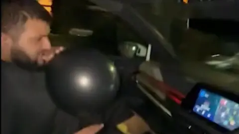 PA Media A still from a video taken on a mobile phone of Usmon Mahmood inhaling from a black balloon. He is in a car being driven by Hussain