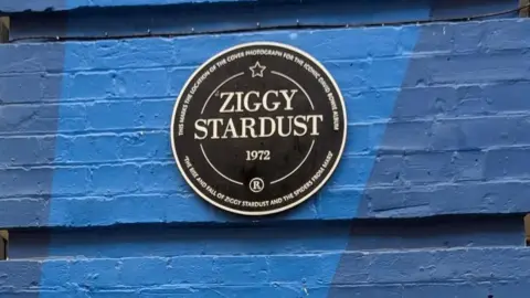 A black circular plaque saying Ziggy Stardust on a blue-painted wall.