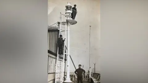 Family handout A black and white imae of Lightships lighthouse in Cornwall. One wearing a captain's hat stands at the top next to the glass light. Orlando Stokes can be seen standing mid way down a ladder with another standing at the bottom. 