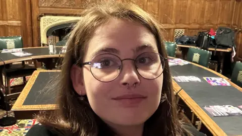 A young woman with brown hair and a septum piercing stands in a room in the House of Commons. Behind her are green leather chairs with gold crown emblems on them, and a long table.