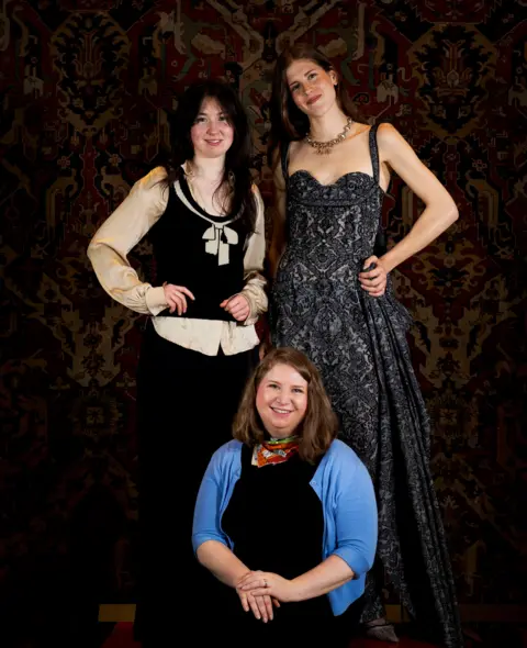 Burrell Collection Three women smile for the camera - two of the women are standing up, Flora McFarlane, with long dark hair, a black and beige top, and Danae Crawford in a long black dress.
Sitting in front of them is Juliet Fellows Smith, wearing a black and blue. She has shoulder length auburn hair.