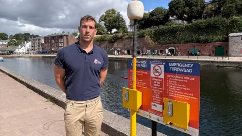 RLSS UK A man wearing a blue polo shirt and brown trousers is stood next to a yellow, red and blue safety sign. Both the man and the sign are stood infront of a river. There are some houses and trees in the background, and the sky is blue with some clouds.