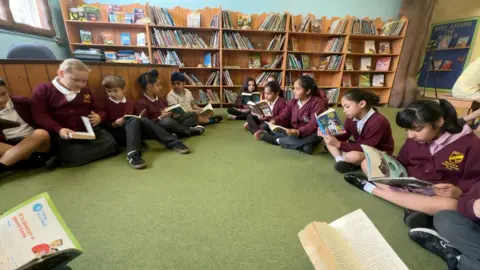 About a dozen children sit on the edge of a classroom while each individually reading books. There is a bookcase in the background filled with books. The carpet is green.