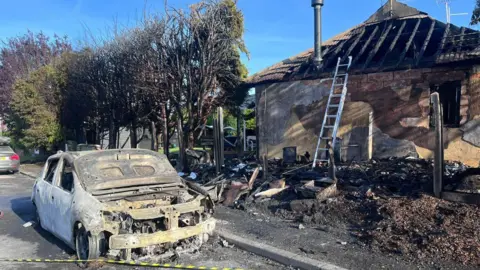 Essex Fire and Rescue Service A burnt out white car, with the bonnet up, is next to the pavement. There is charred debris and trees next to a building which has been badly damaged, with the roof beams visible. 