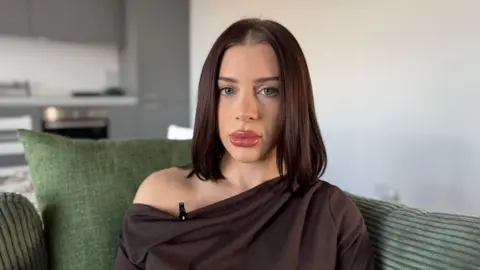 Charlie Rose/BBC A woman sat on a green sofa. A blank wall and a kitchen can be seen in the background.