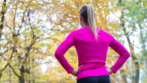 A woman runner standing still with her back to the camera, wearing a bright pink top, standing with her hands on her hips. She has long blonde hair in a pony tail and there are autumnal leaves on the trees.