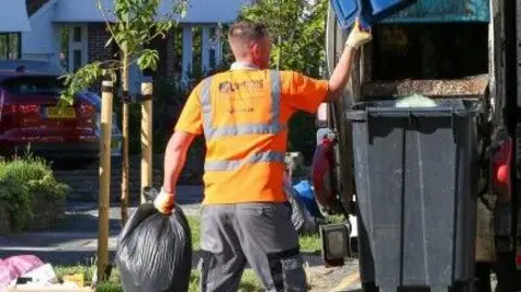 LDRS A waste and refuse collector holding a bin bag and putting rubbish into a black wheelie bin.