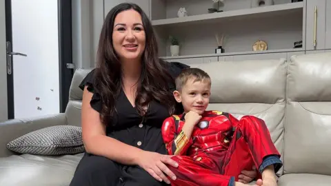 Jasmin with long black hair wearing a black dress cuddles Isaac who has short brown hair and is wearing a red Iron Man superhero outfit. They are sat on a grey sofa with shelving behind it 
