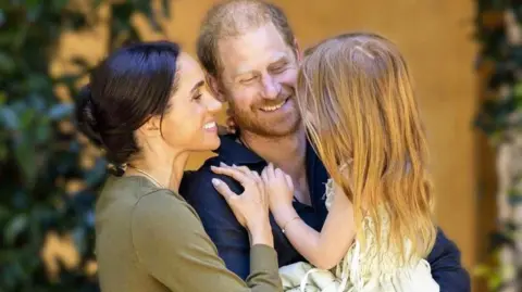 Duke and Duchess of Sussex in casual outfits embracing and holding Lilibet in a summer dress, she has bare feet and her back is to the camera; she has long, red hair