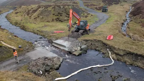 Tees River Trust Aerial view of work to remove a ford over a beck. There is a gap in the narrow road across it, with a digger on one side and a workman in high viz uniform standing on the other. The road winds back across rolling moorland.