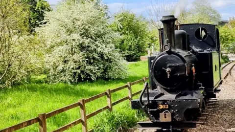 BBC Weather Watchers/Stevo100 A black steam engine rides towards us, alongside a fence and trees.