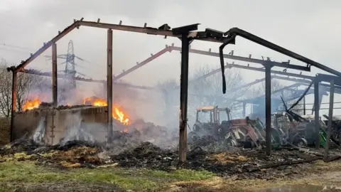 HIWFRS The aftermath of the fire, including hay burning on the left hand side and three wrecked tractors, with just the barn's metal frame left after the blaze