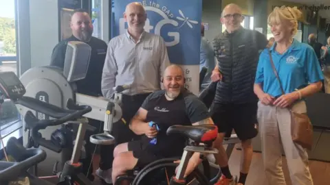 Mike, who is in his wheelchair, smiling while he holds his medal. He wears a black T-shirt and has three men and a woman standing around him in front of a Bridge The Gap banner.