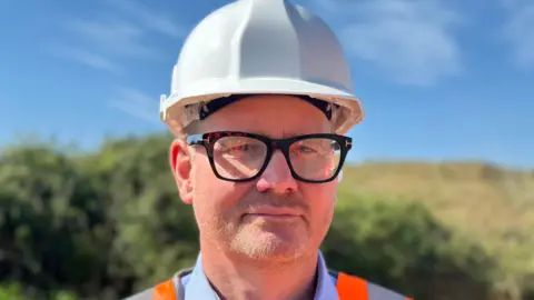 David Rice has black glasses and is wearing a white hard hat, blue shirt and orange hi-vis jacket. Behind him are blurred green bushes and a brown landfill.