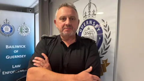 BBC Damien Kitchen, a middle aged man dressed in a black police uniform with his arms crossed. He is standing in front of a wall with the Guernsey Police logo in the background 