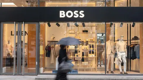 The entrance of the Hugo Boss flagship fashion store in Paris, France. In the foreground, a blurred woman is walking past with an umbrella. Inside the shop, mannequins, shirts, shoes, and trousers are visible.