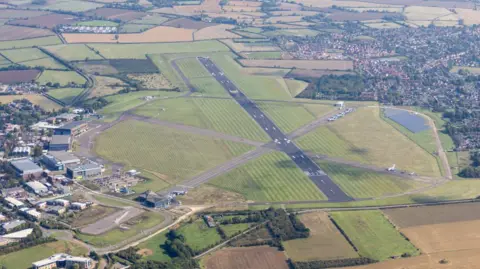 Getty Images An aerial view of Cranfield Airport with the runway in the centre and buildings to the left. There is housing to the right with green fields at the top and bottom of the photo.